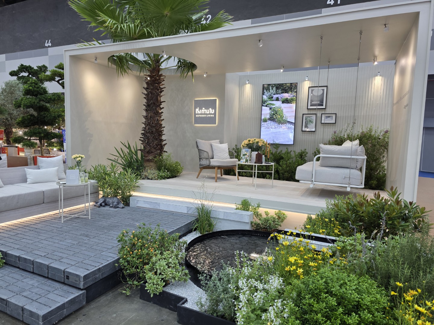 A stylish outdoor living booth featuring cozy seating, lush plants, and a serene pond