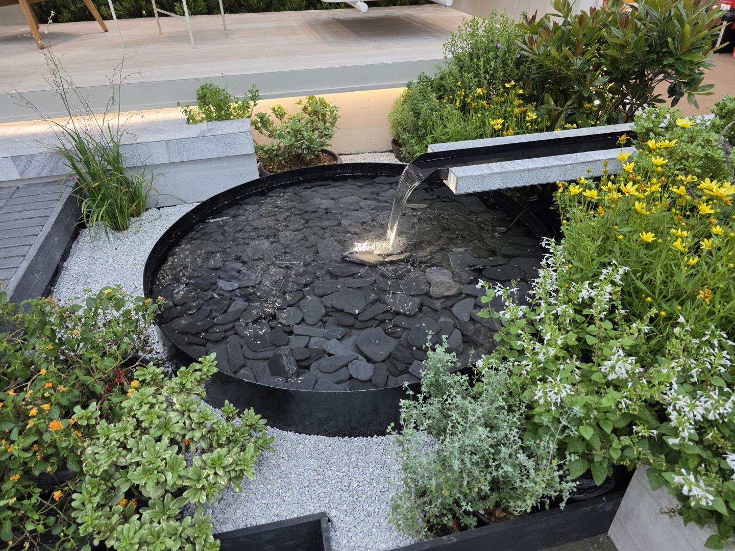 A modern garden pond with a sleek stone water feature, surrounded by vibrant flowers and greenery.