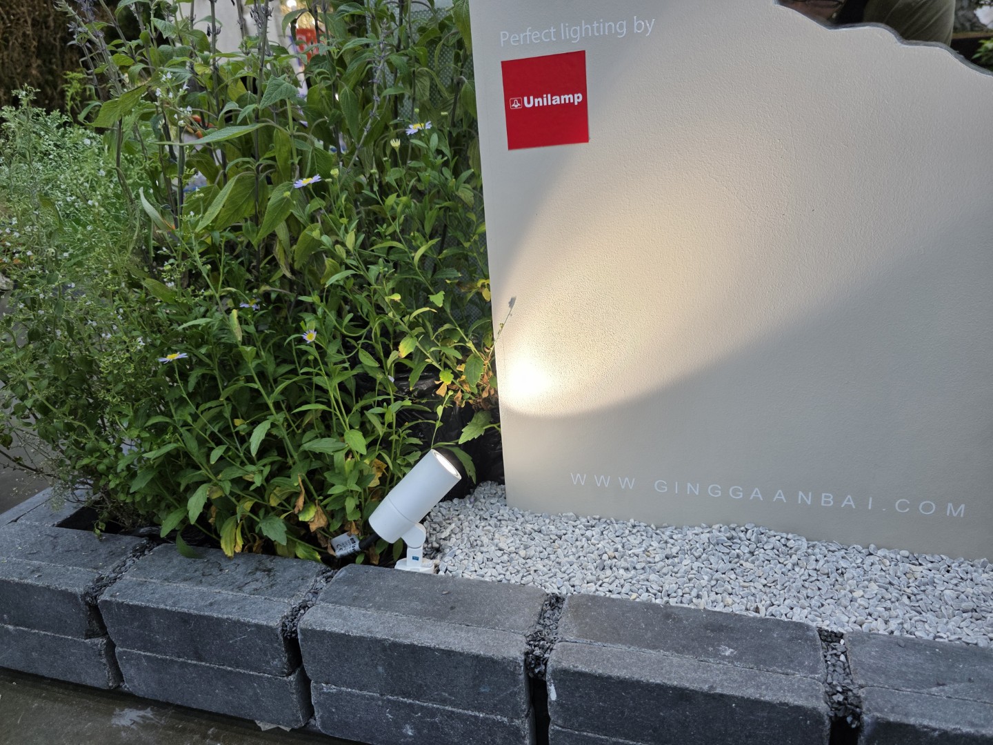 A spotlight from Unilamp illuminates a garden sign surrounded by lush plants and white gravel.