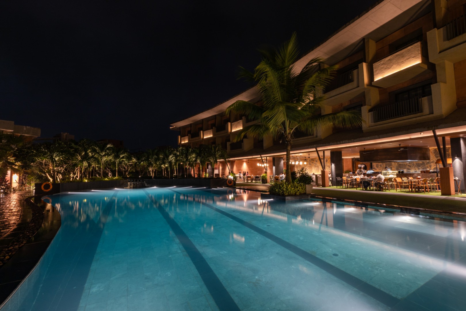 Illuminated resort swimming pool with palm trees and open-air restaurant at night.